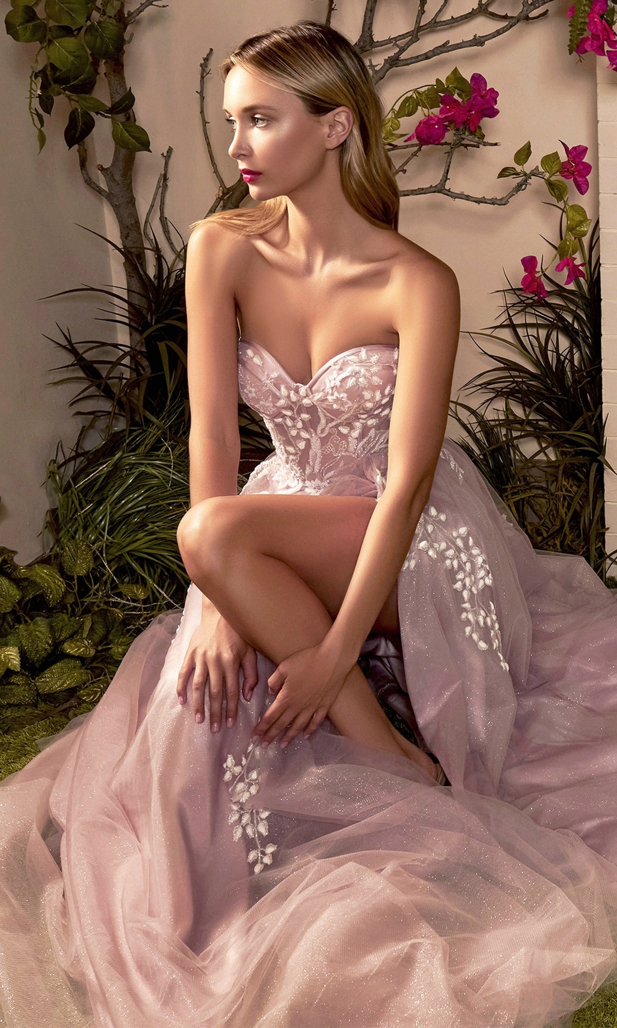 A woman wearing the Strapless Sweetheart Prom Ball Gown A1029 with floral embroidery sits amidst greenery and pink flowers, one knee raised as she gazes to the side.